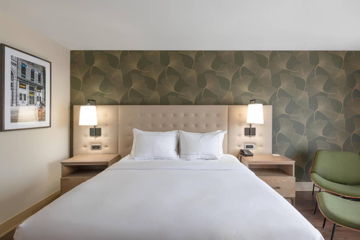 A hotel room with a large bed, two bedside lamps, a green chair, and a wall with a leafy pattern.