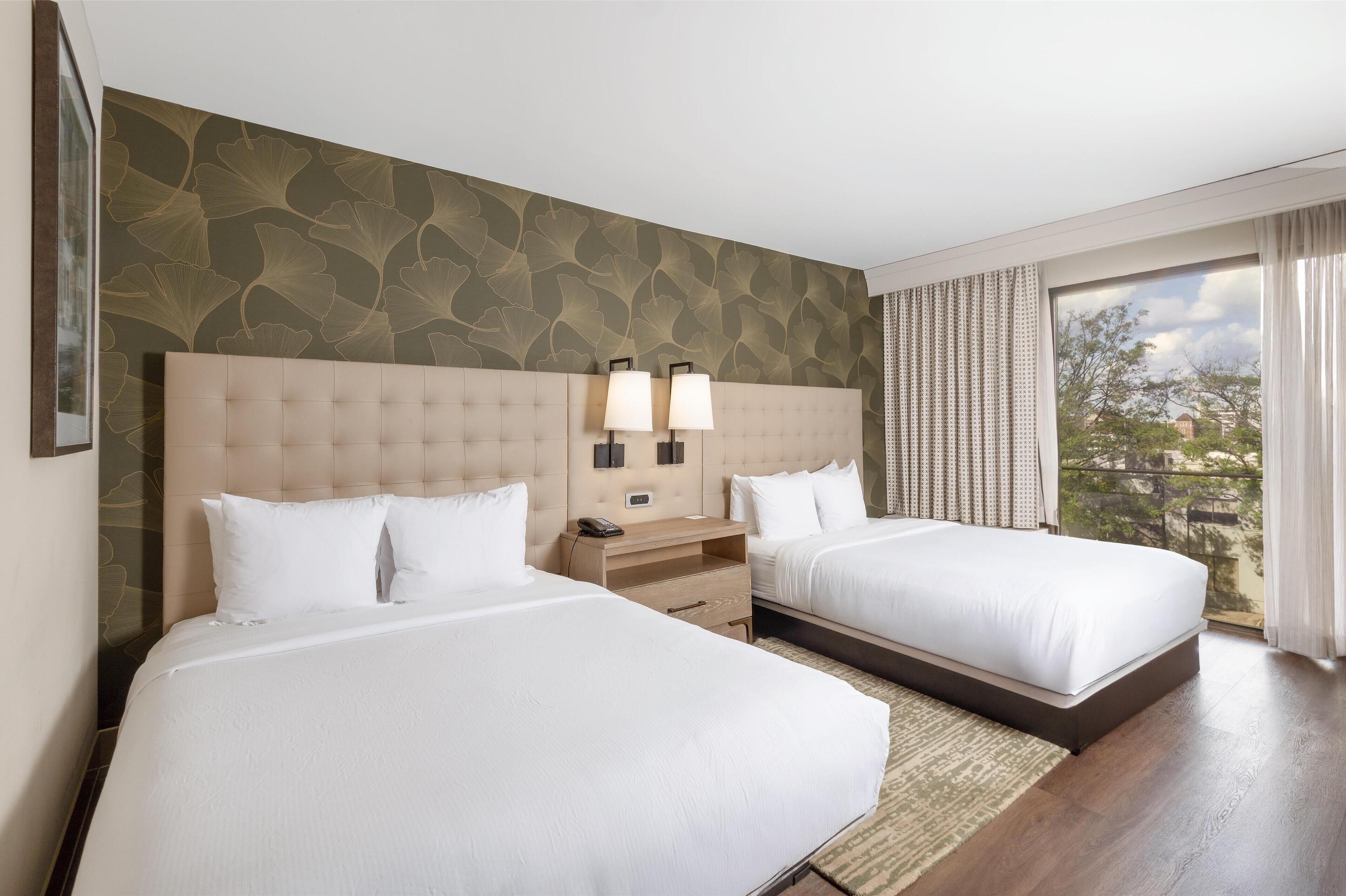 A hotel room with two beds, a large window with curtains, a wall with a leafy pattern, and a bedside table with lamps.