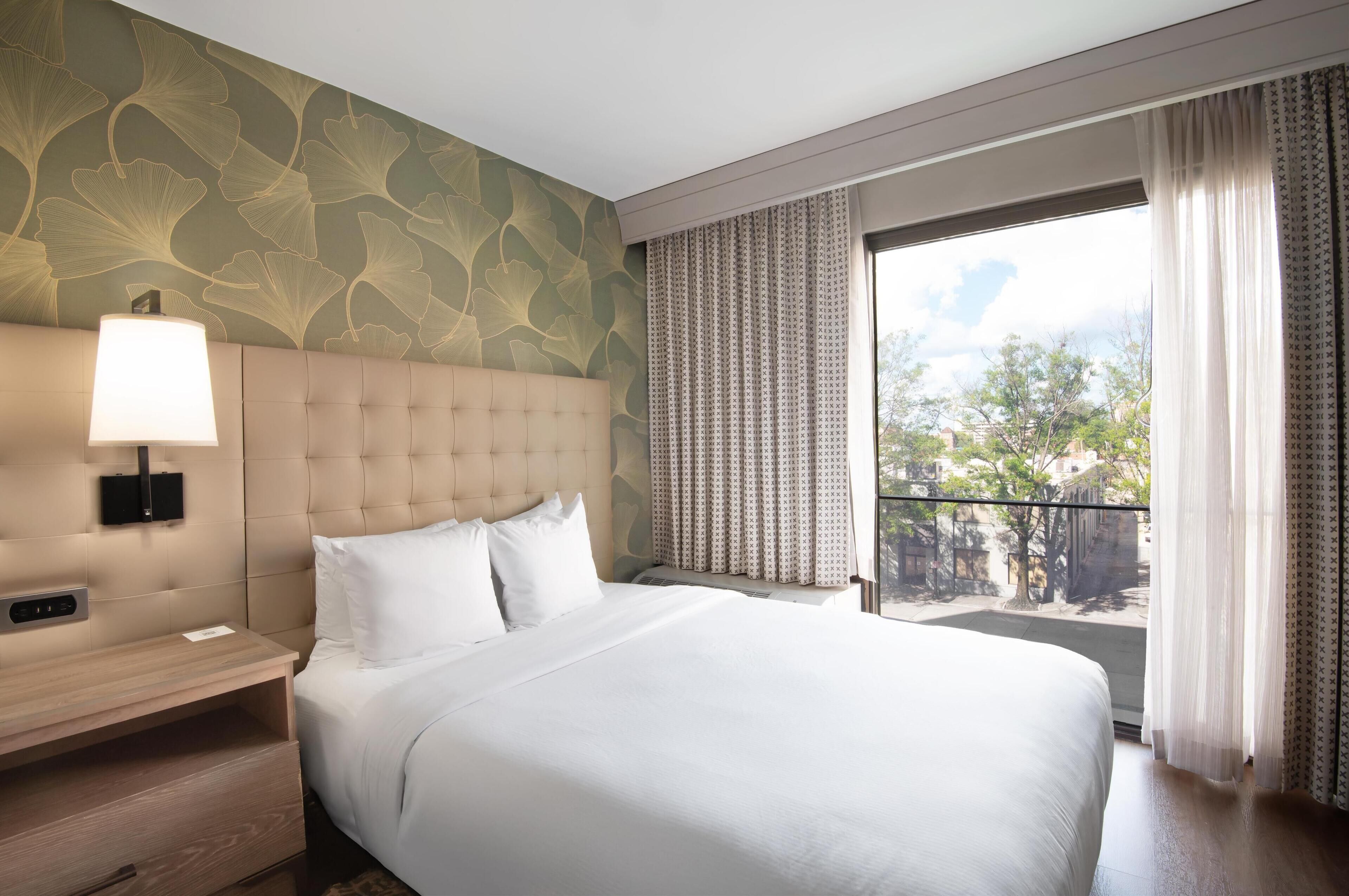 A hotel room with a large bed, a headboard with a light fixture, a bedside table, and a window with curtains.