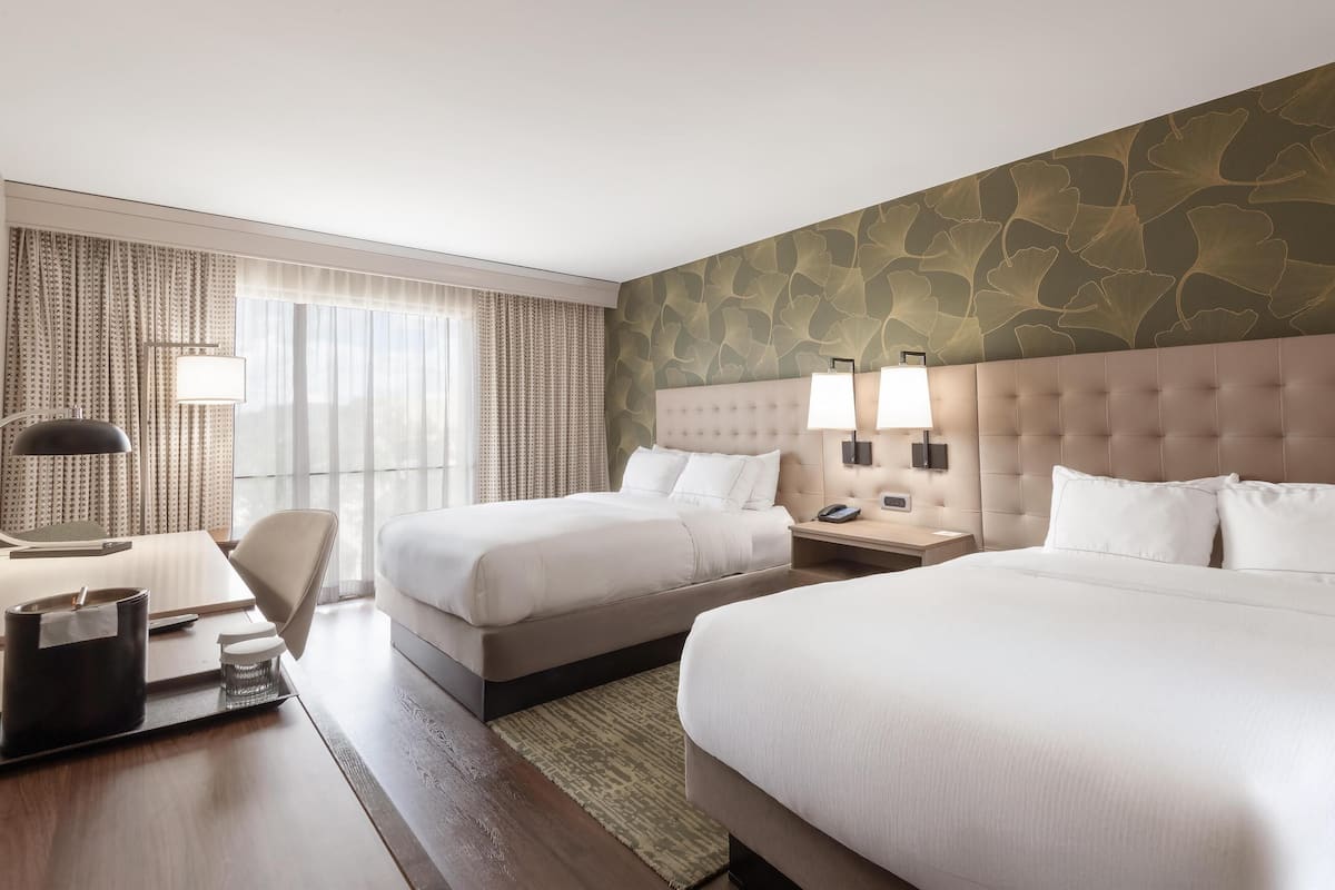 A hotel room with two beds, a desk, and a large window with curtains.