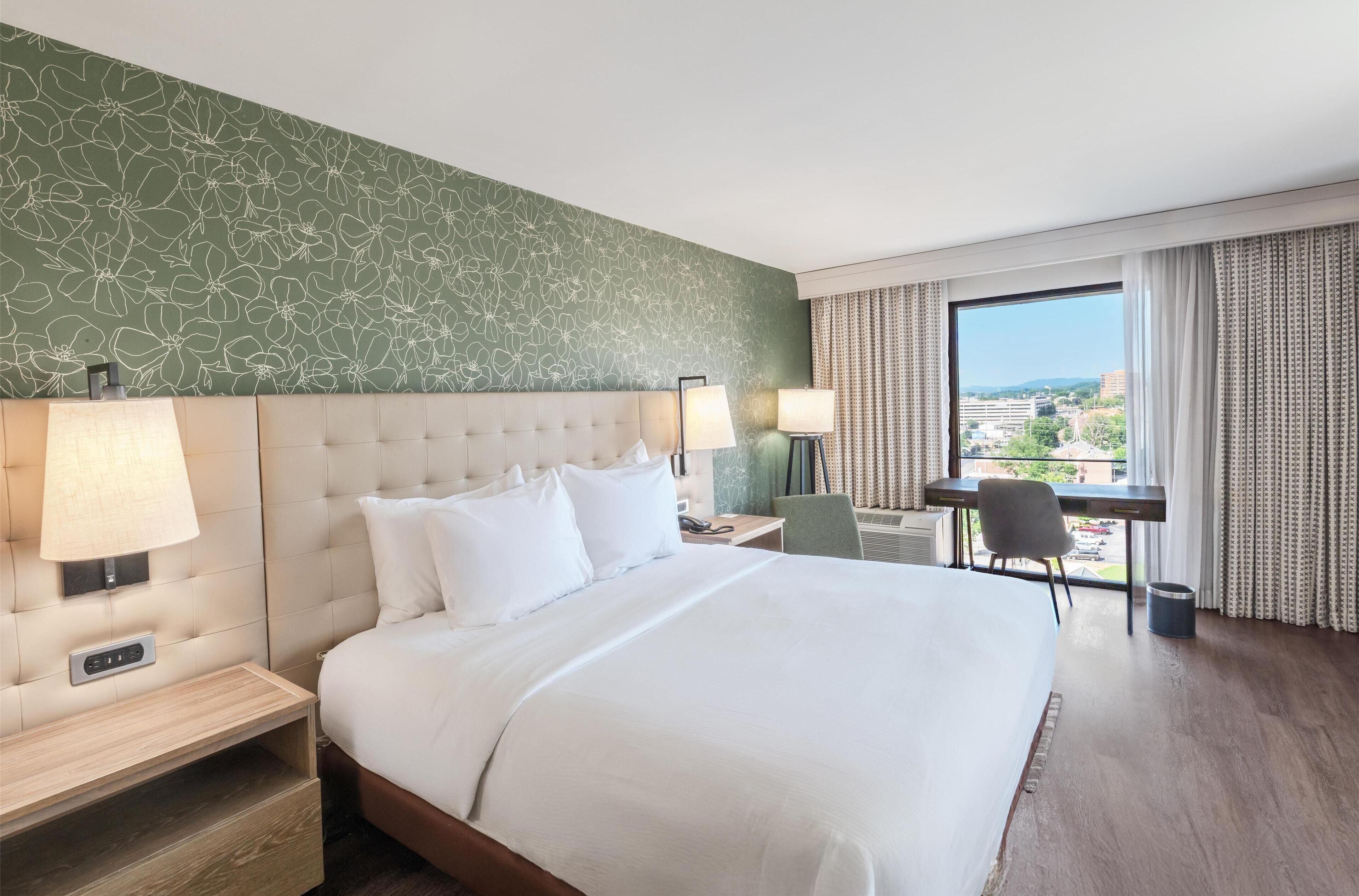 Une chambre d’hôtel avec un grand lit, deux lampes de chevet, un bureau et une vue sur la ville par une grande fenêtre.