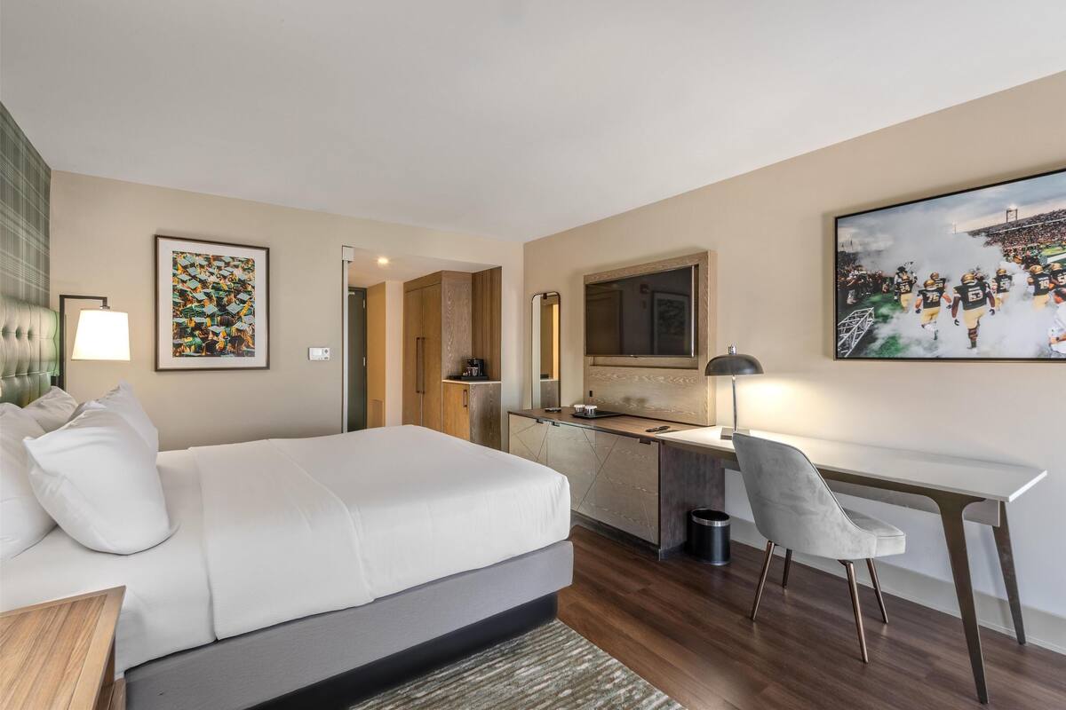 A modern hotel room with a large bed, a desk, a chair, a mirror, and a television.