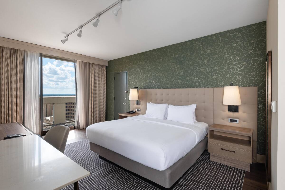 A hotel room with a large bed, a desk, a chair, a nightstand with a lamp, and a view of the city.