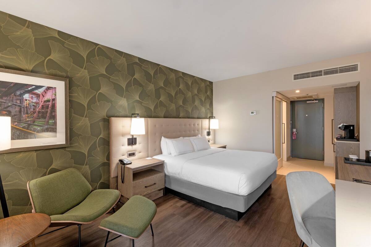 A hotel room with a bed, two chairs, a desk, and a framed picture on the wall.