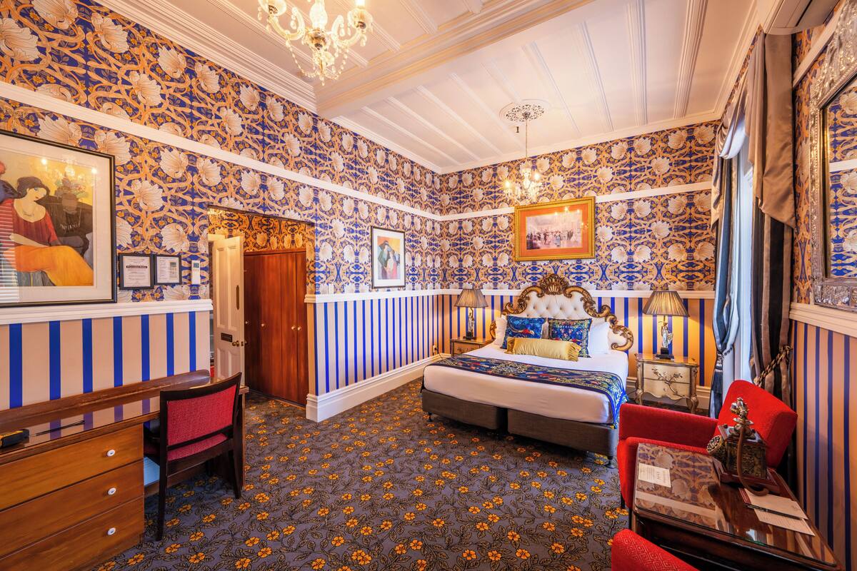 Un dormitorio con papel tapiz estampado, una cama con cabecera, un escritorio con silla y un sillón rojo.