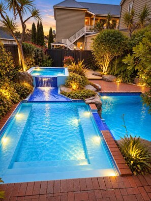 Una piscina rectangular con cascada, rodeada por un jardín ajardinado con diversas plantas y una casa al fondo.
