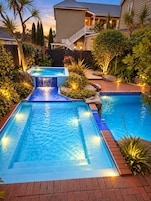 A rectangular swimming pool with a waterfall feature, surrounded by a landscaped garden with various plants and a house in the background.