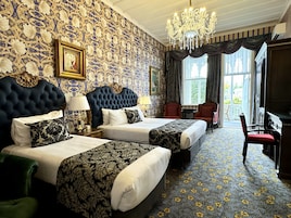 A hotel room with two beds, a chandelier, a large window with curtains, and a view of the outside.