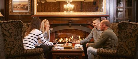 A group of people seated around a table in a room with a fireplace, patterned furniture, and a mirror above the fireplace.