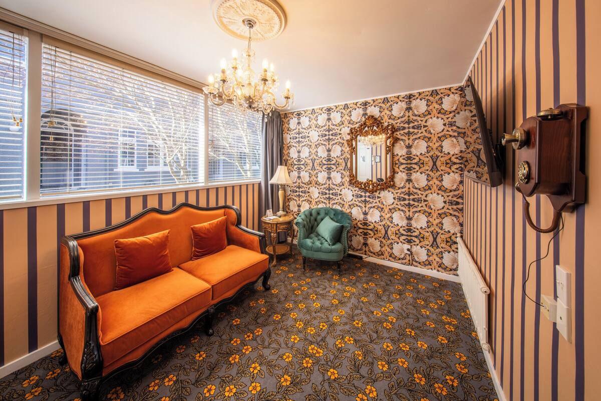 Una habitación con pared a rayas, alfombra estampada, sofá antiguo, sillón verde, candelabro y teléfono.