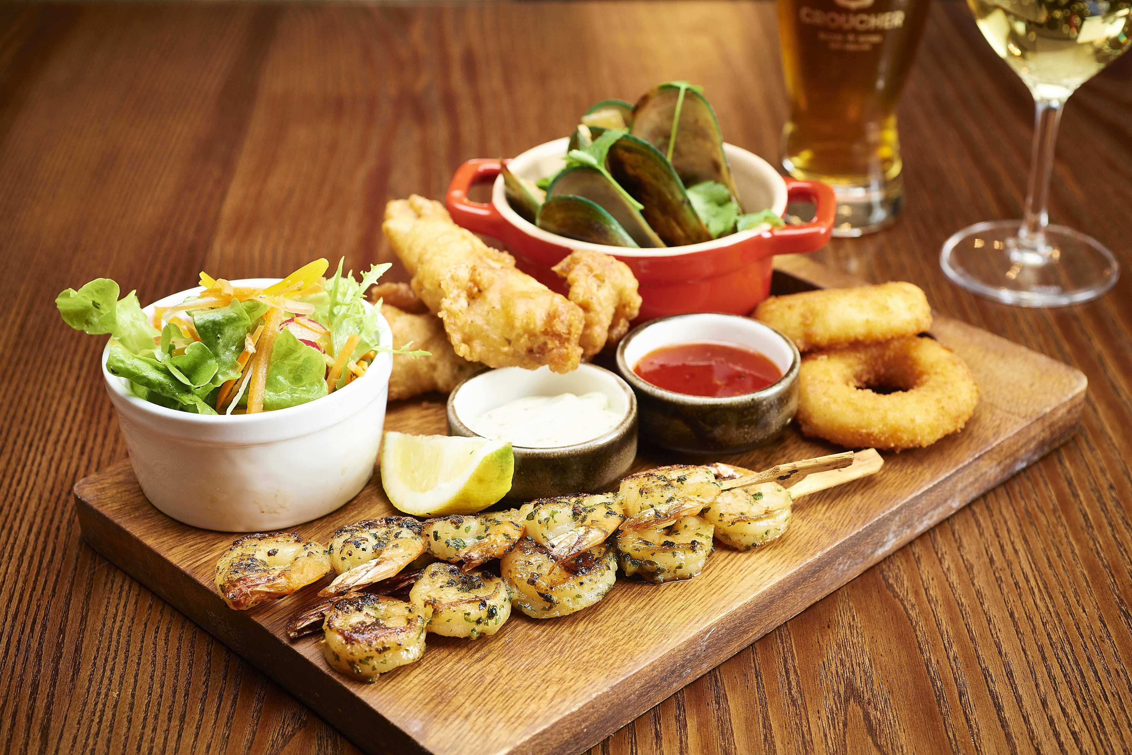 Un plateau en bois sur lequel sont disposés divers plats, notamment des crevettes grillées, des aliments frits et une salade.