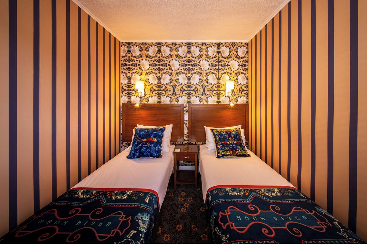Habitación de hotel con dos camas, papel tapiz a rayas, cabecero estampado y almohadones decorativos.