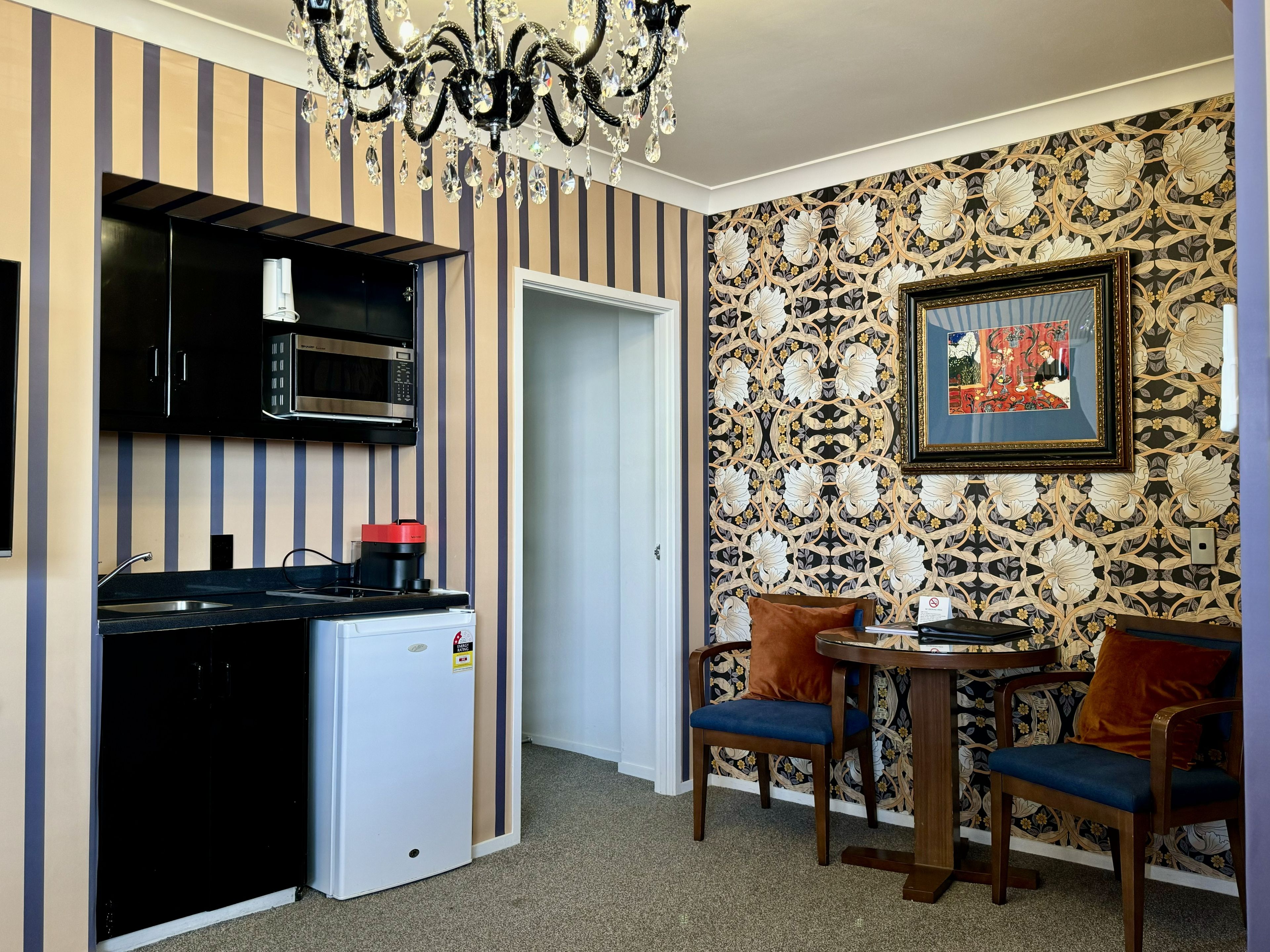 A compact kitchen with a microwave, coffee maker, and a refrigerator. There is a dining area with two chairs and a table, and a chandelier above.