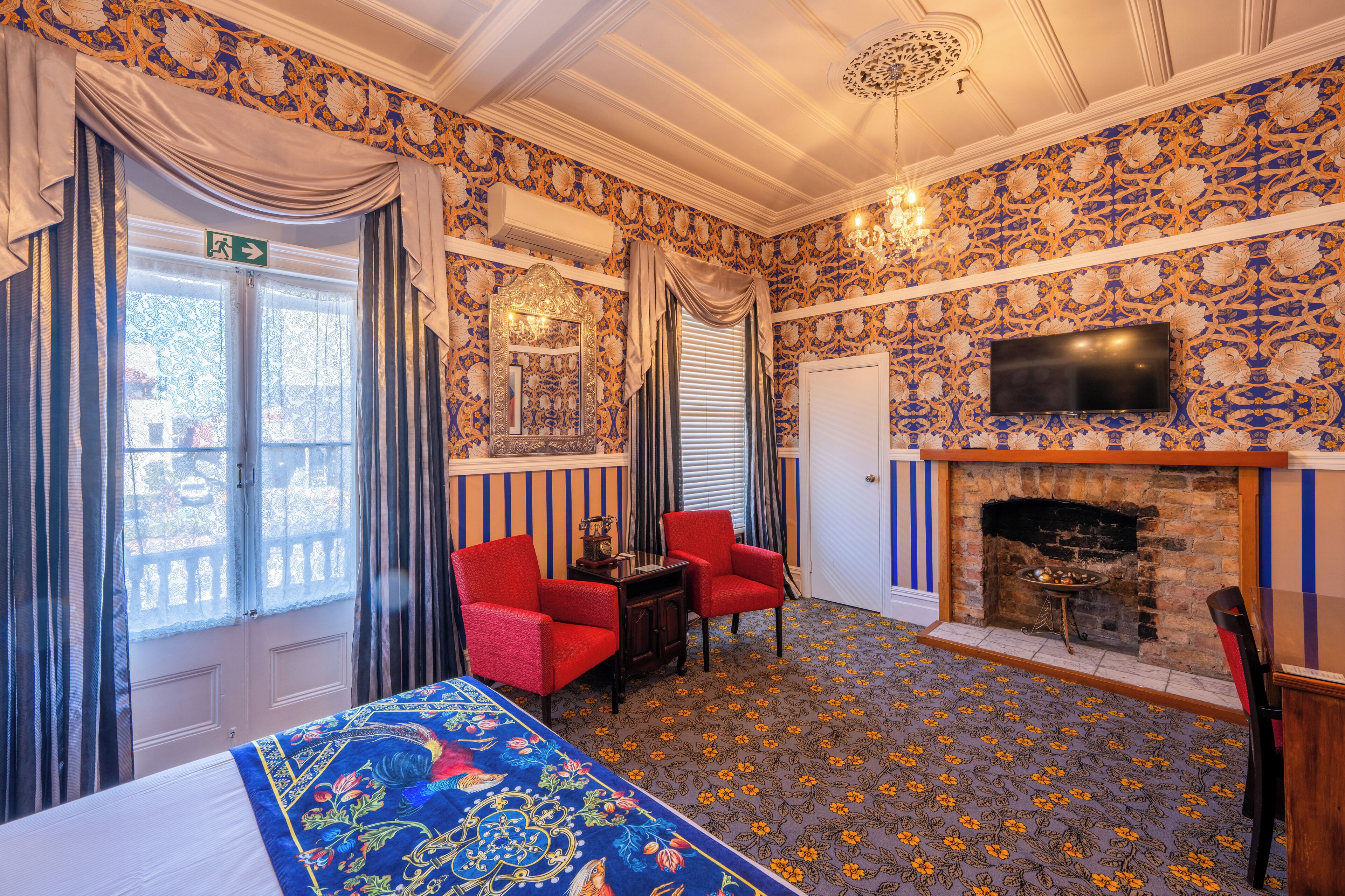 A room with a bed, two red chairs, a fireplace, and a television on the wall.
