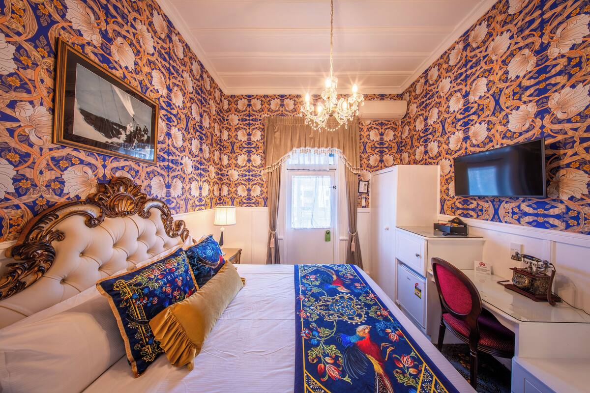 Un dormitorio con cabecera capitonada, una cama grande con almohadas decorativas, un escritorio con silla, un televisor y una araña.