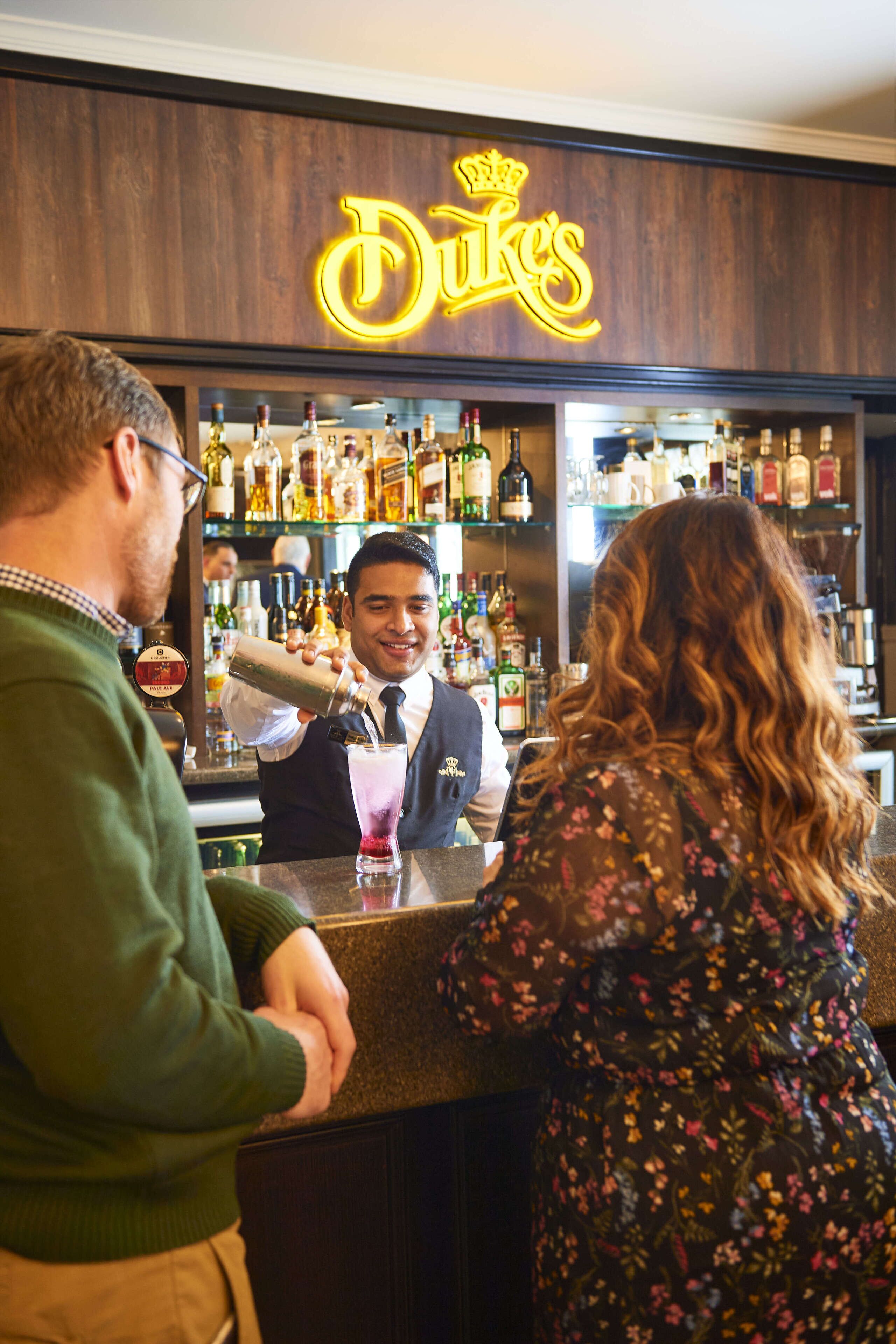 En bar med en neonreklamskylt som visar ordet ”Dukes”, och en bartender som gör i ordning en drink.