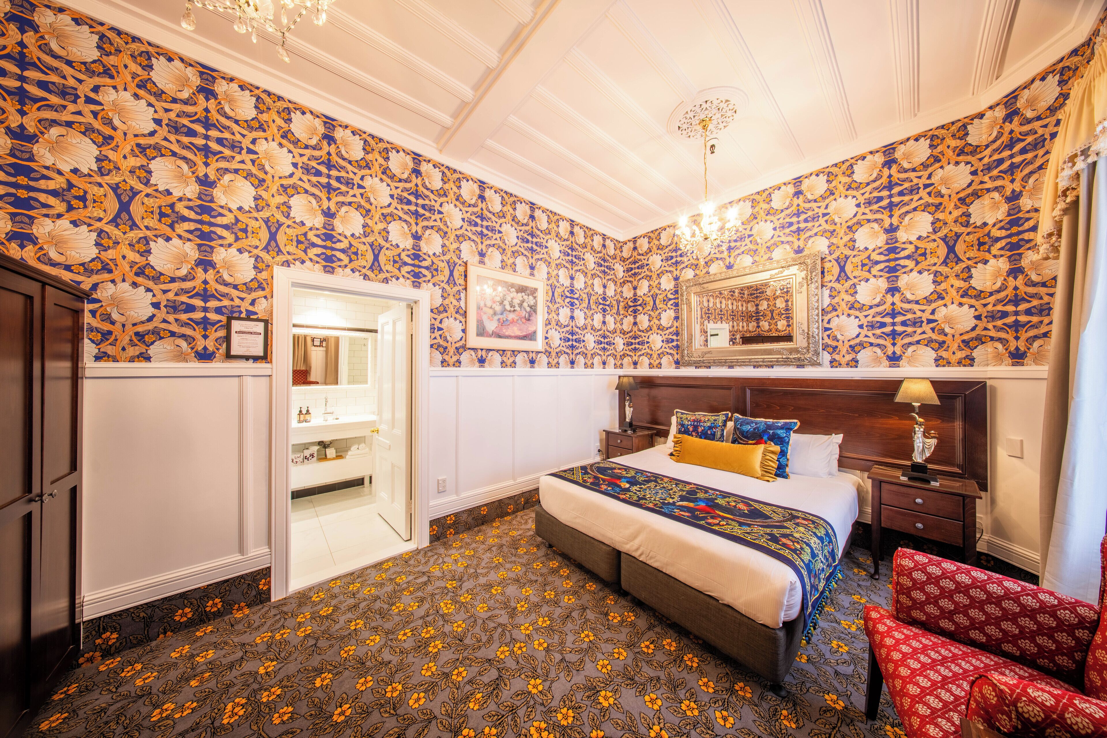 Una habitación de hotel con papel tapiz estampado con motivos florales, una cama grande con colcha estampada, una mesita de noche con lámpara y un sillón rojo.