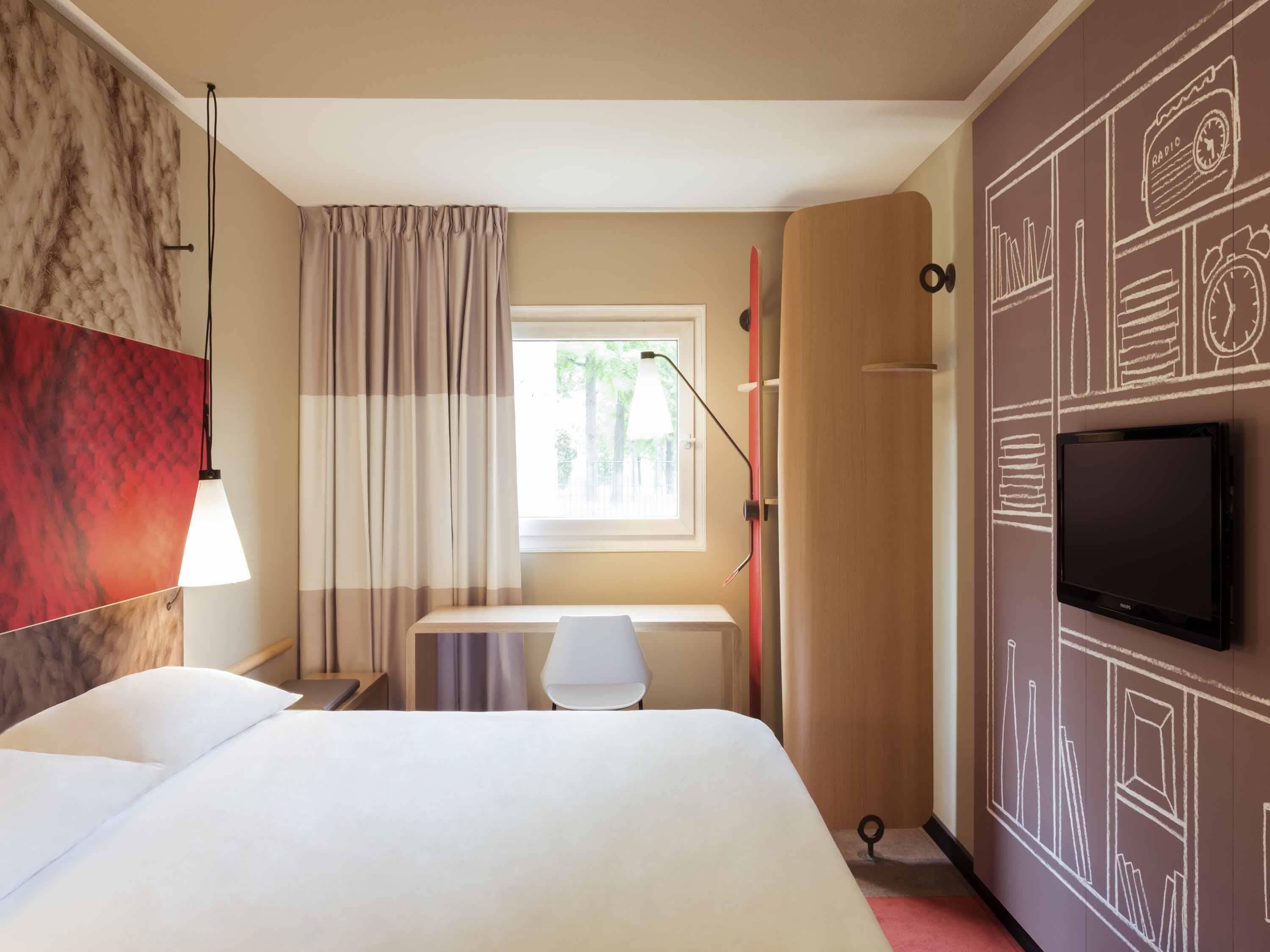 Une chambre d’hôtel avec un lit, un bureau, une télévision, une fenêtre avec des rideaux et un mur orné d’une horloge et d’une radio.