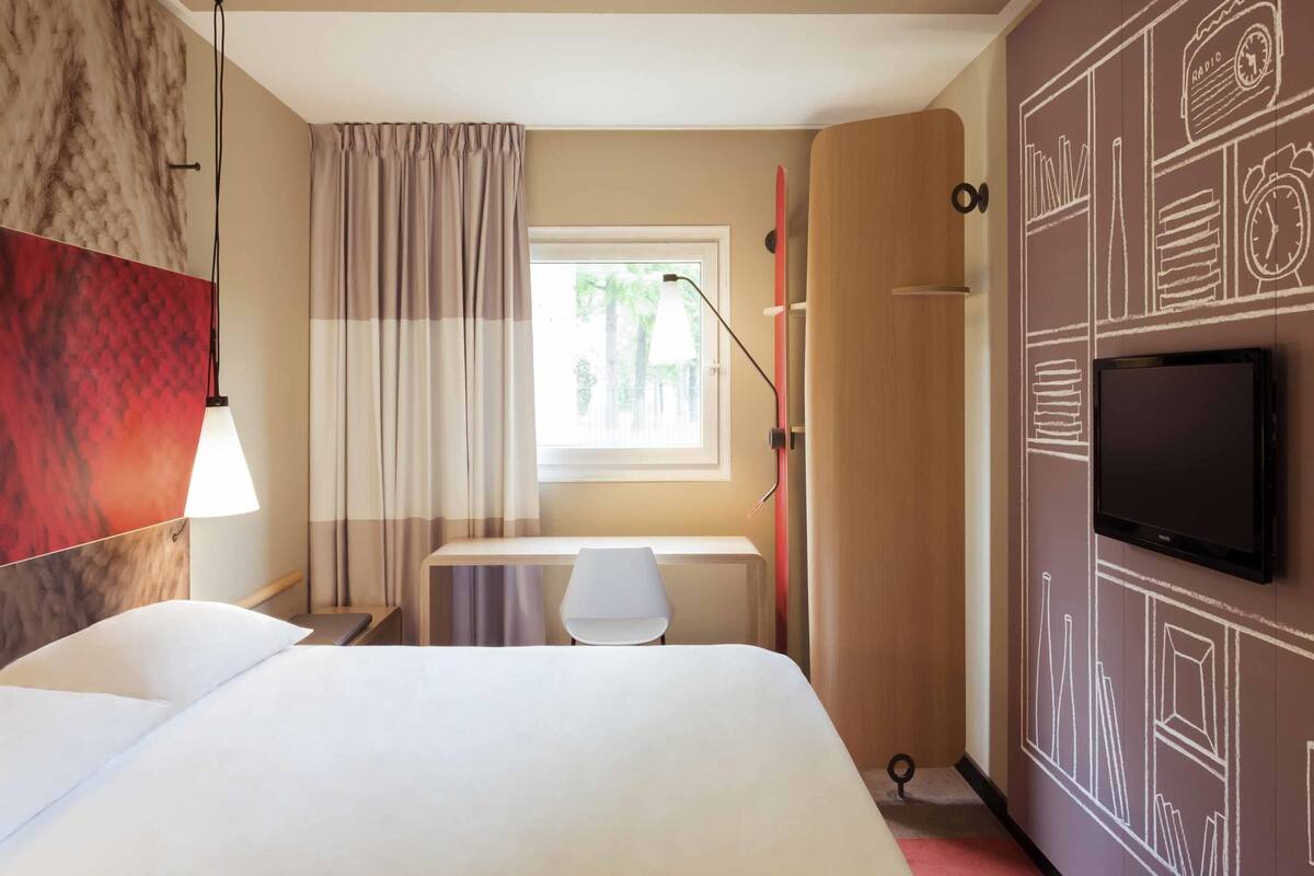 A hotel room with a bed, a desk, a TV, a window with curtains, and a wall with a clock and radio.