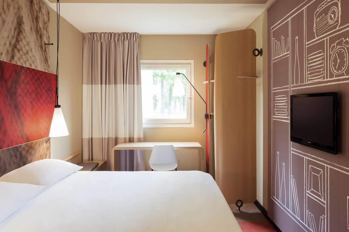 A hotel room with a bed, a desk, a TV, a window with curtains, and a wall with a clock and radio.