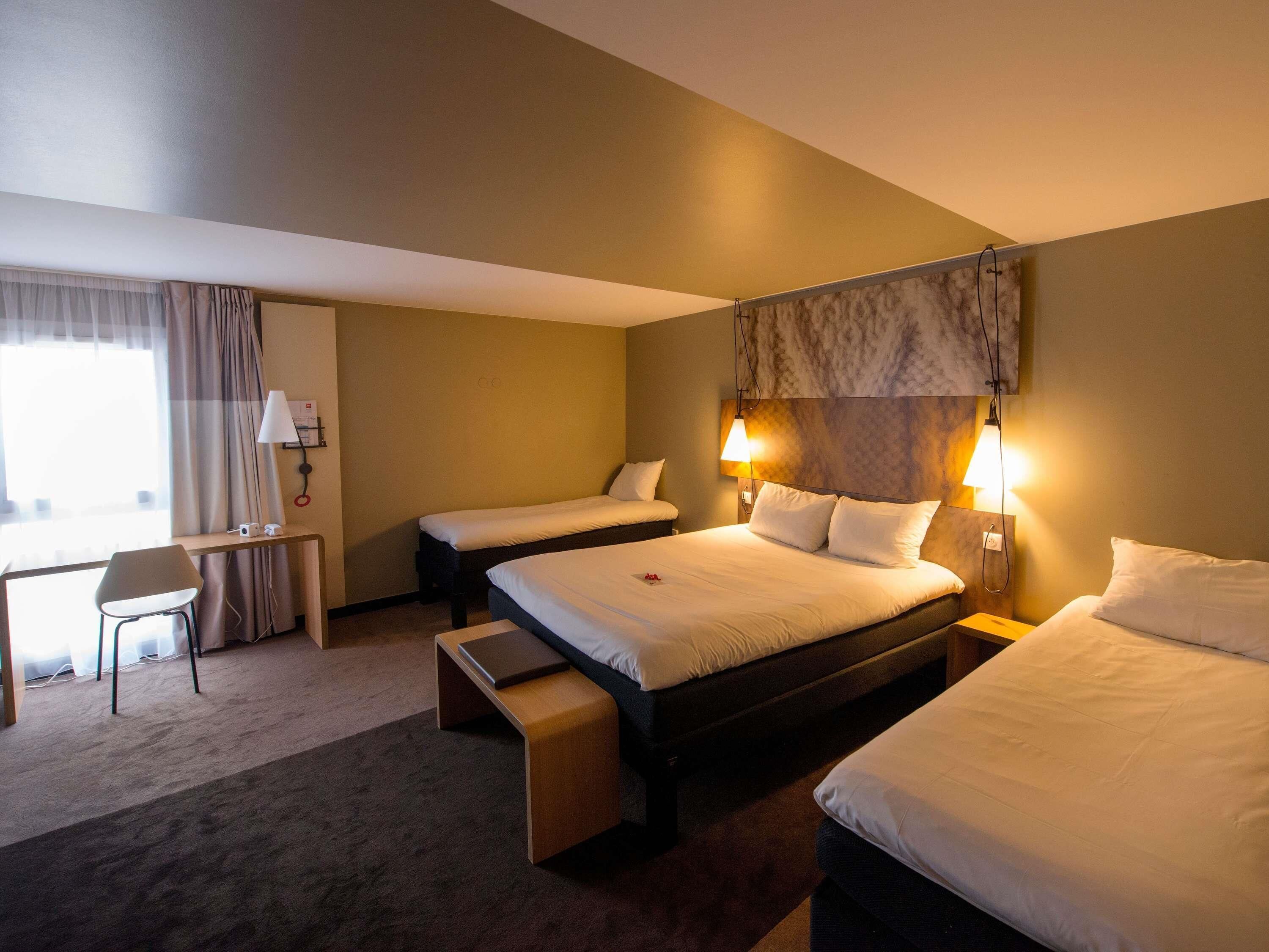 Quarto de hotel com duas camas, uma escrivaninha, uma cadeira e um pequeno banco.