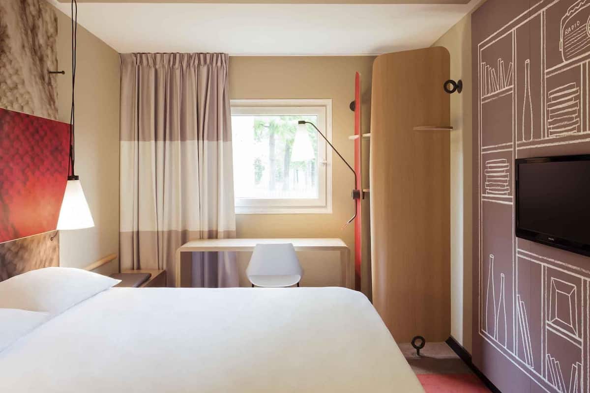 Una habitación de hotel con cama, escritorio, televisor, ventana con cortinas y una pared con diseño geométrico.