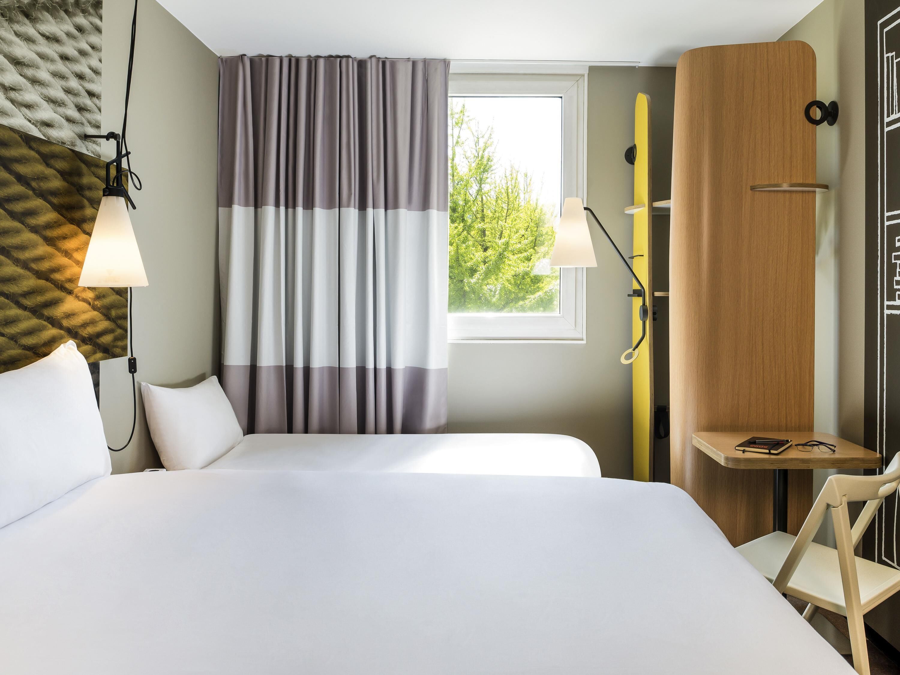 A hotel room with a bed, two bedside lamps, a window with curtains, a wooden wardrobe, and a small table with a chair.