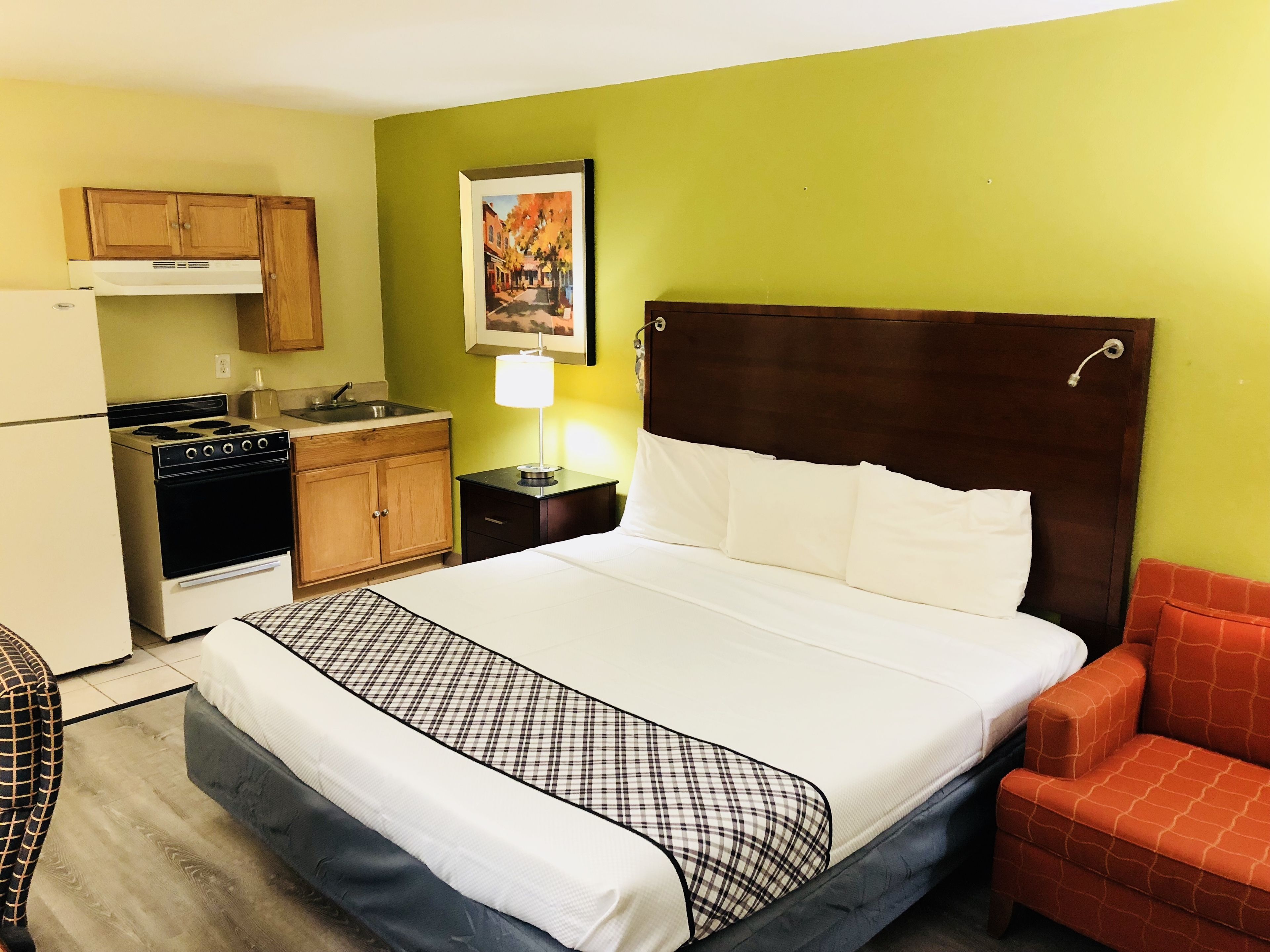 A hotel room with a bed, a nightstand, a lamp, a chair, a kitchen area with a stove, and a refrigerator.