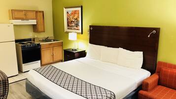 A hotel room with a bed, a nightstand, a lamp, a chair, a kitchen area with a stove, and a refrigerator.