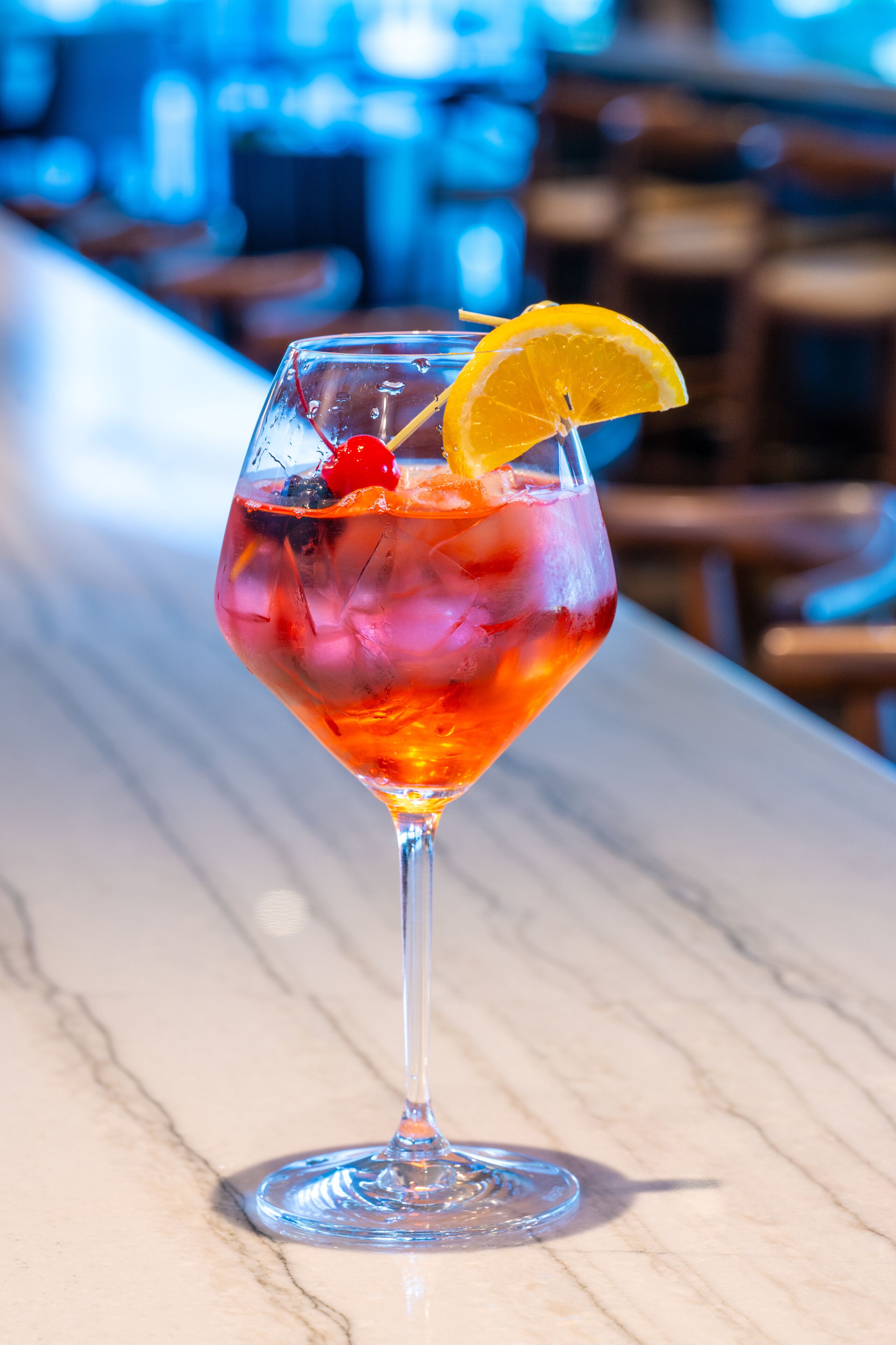 A cocktail in a stemmed glass with ice cubes, garnished with a slice of orange and a cherry, on a marble bar counter.