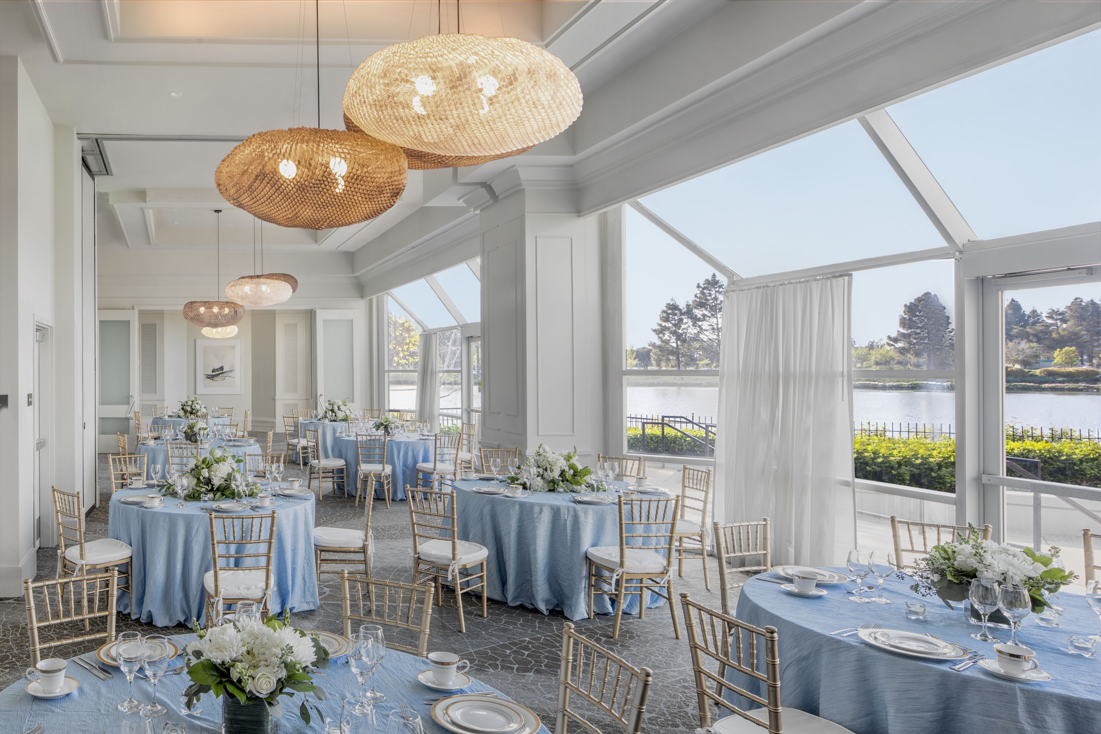 A restaurant with round tables set for dining, large windows offering views of a body of water, and hanging wicker lamps.