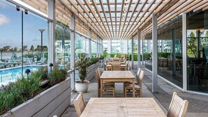 An outdoor patio with wooden tables and chairs, a pool, and glass walls offering views of the surrounding area. - Grand Bay Hotel San Francisco (Redwood City)