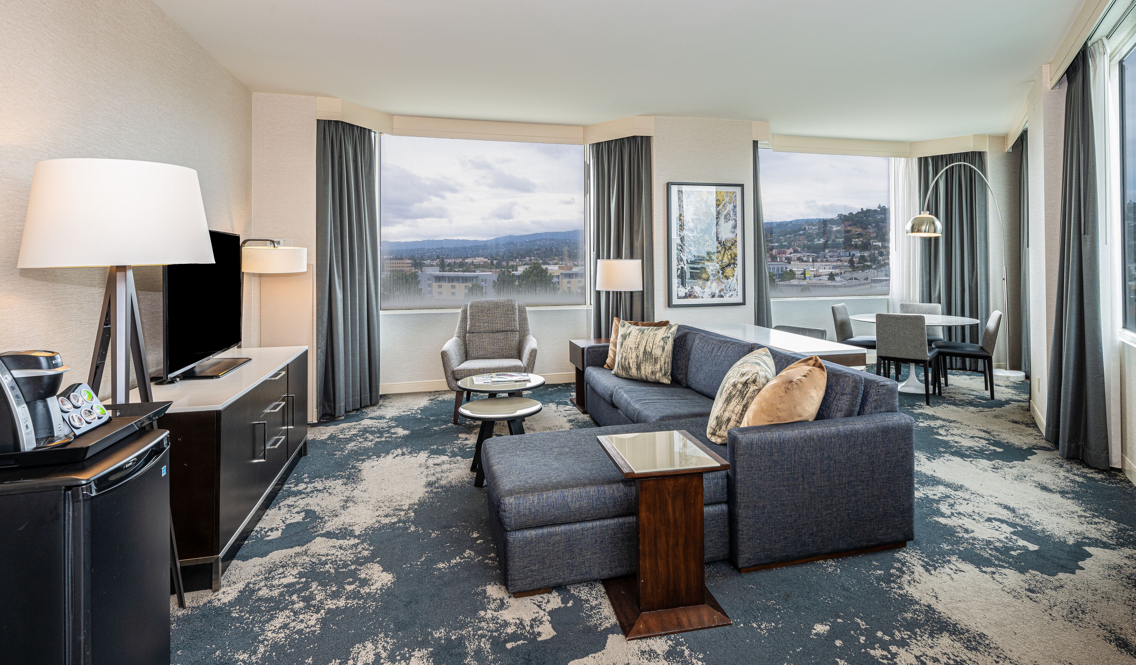 A modern hotel room with a large window offering a city view, a sofa, a small table, a coffee machine, and a TV.