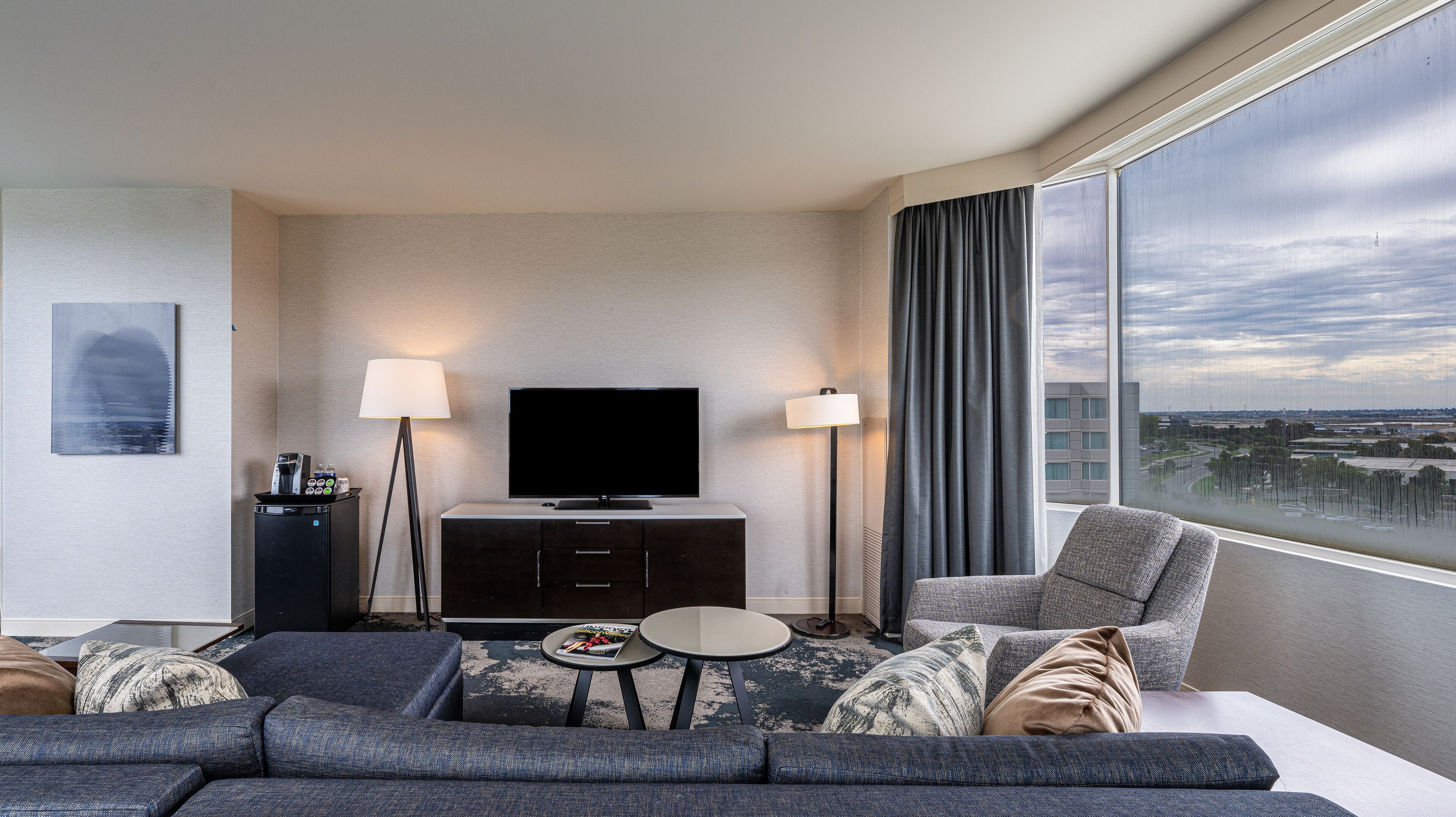 A modern hotel room with a large window, a flat-screen TV, a sofa, a coffee table, and a side table.