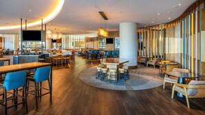 A modern hotel lobby with a curved ceiling, wooden flooring, and a mix of seating areas including bar stools and cushioned chairs. - Grand Bay Hotel San Francisco (Redwood City)