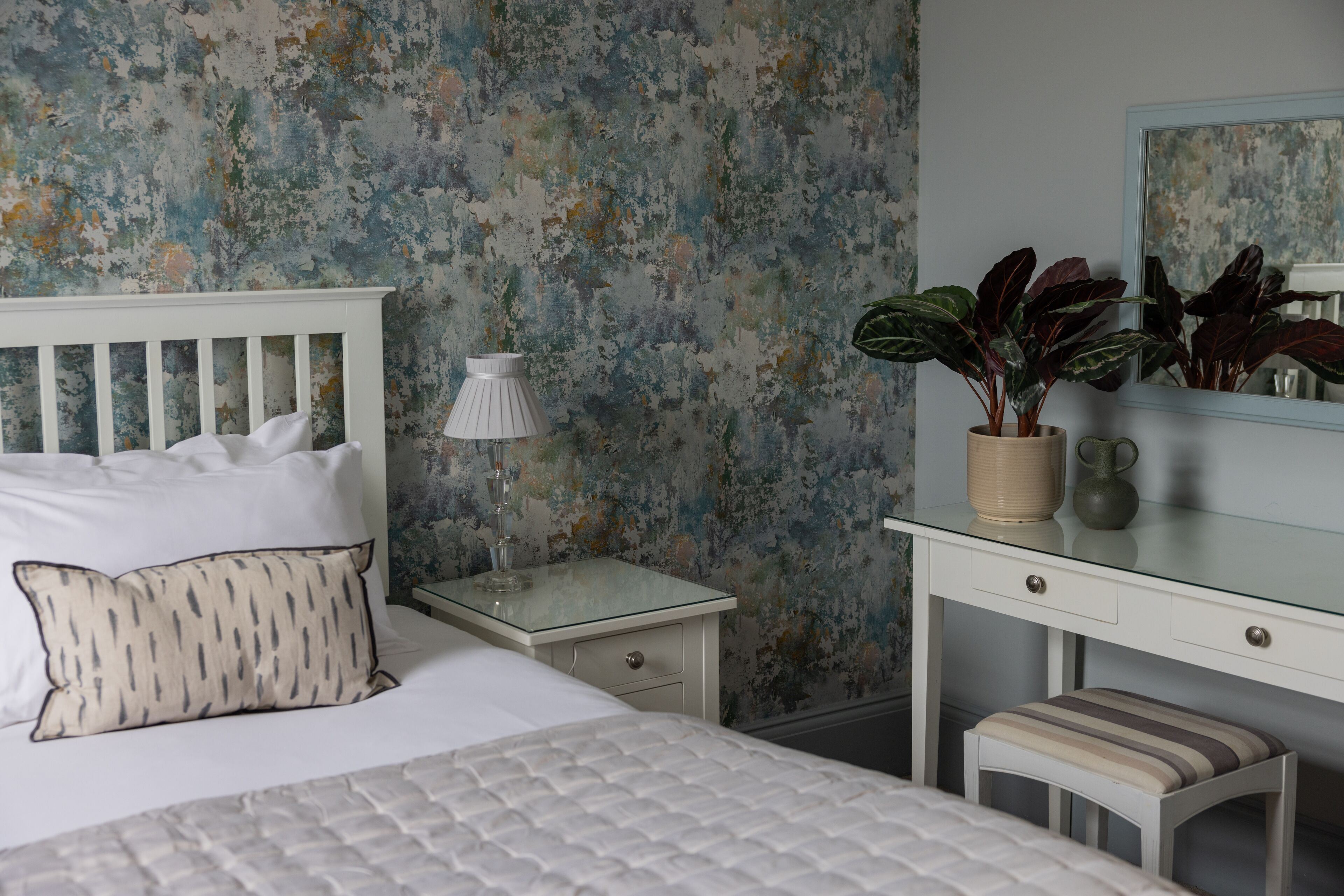 Un dormitorio con cama blanca, lámpara de noche, mesita de noche, cómoda, espejo y una planta en maceta.
