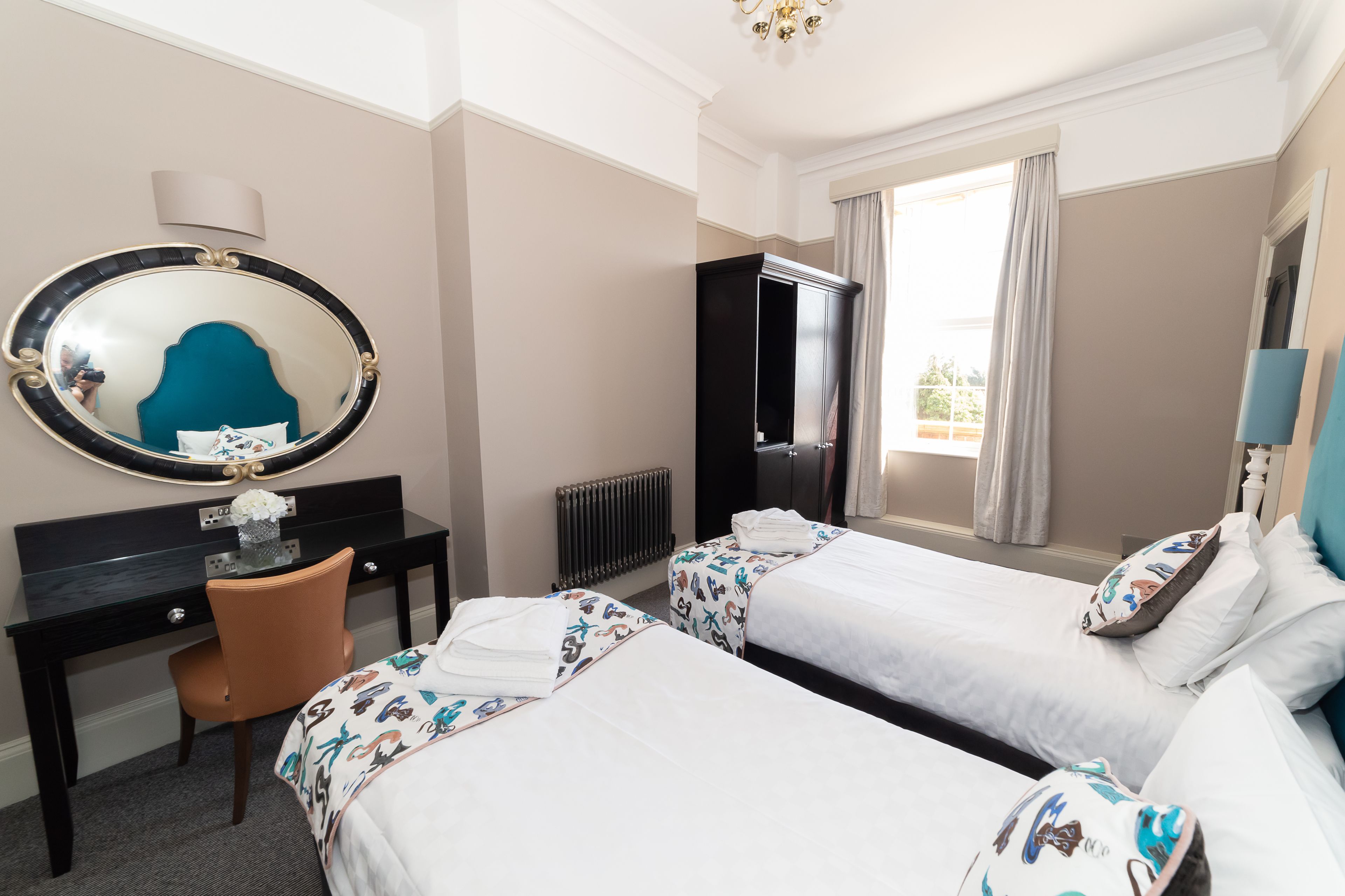 A hotel room with two beds, a desk, a chair, a large mirror, a wardrobe, and a window with curtains.