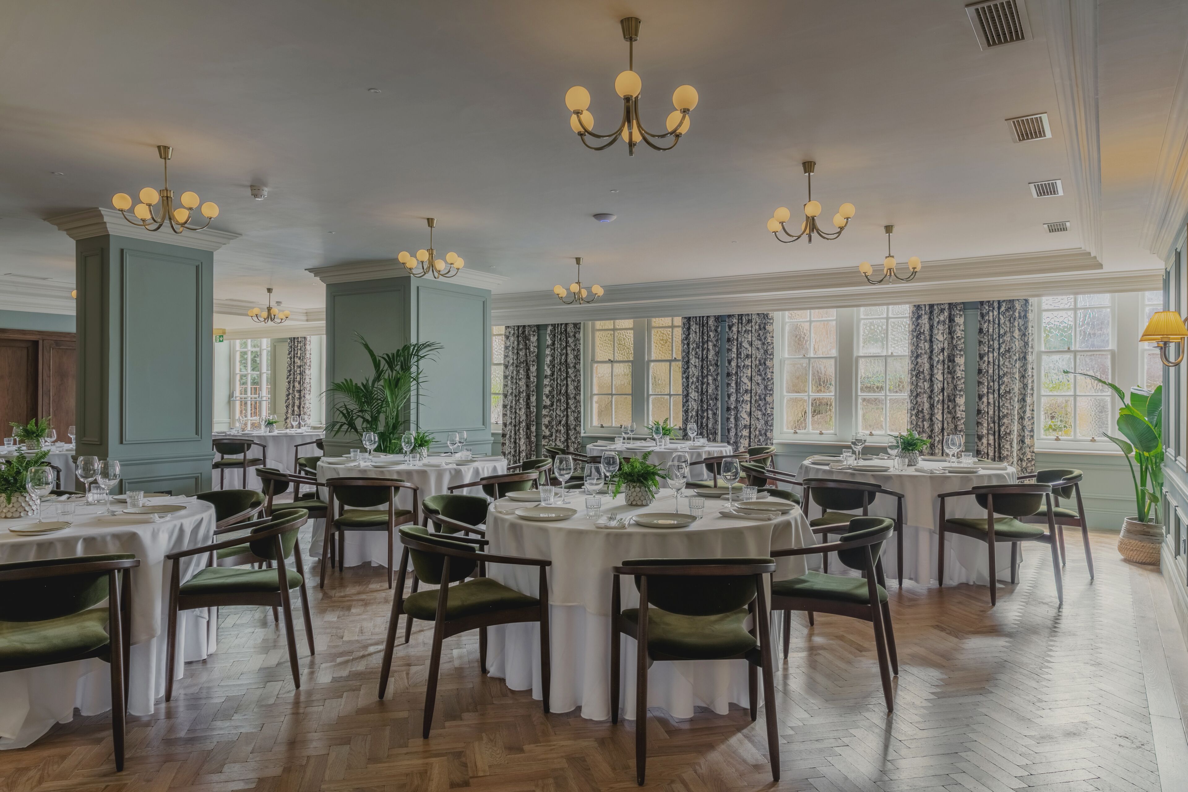 A formal dining room with multiple round tables set for service, green upholstered chairs, and classic chandeliers.