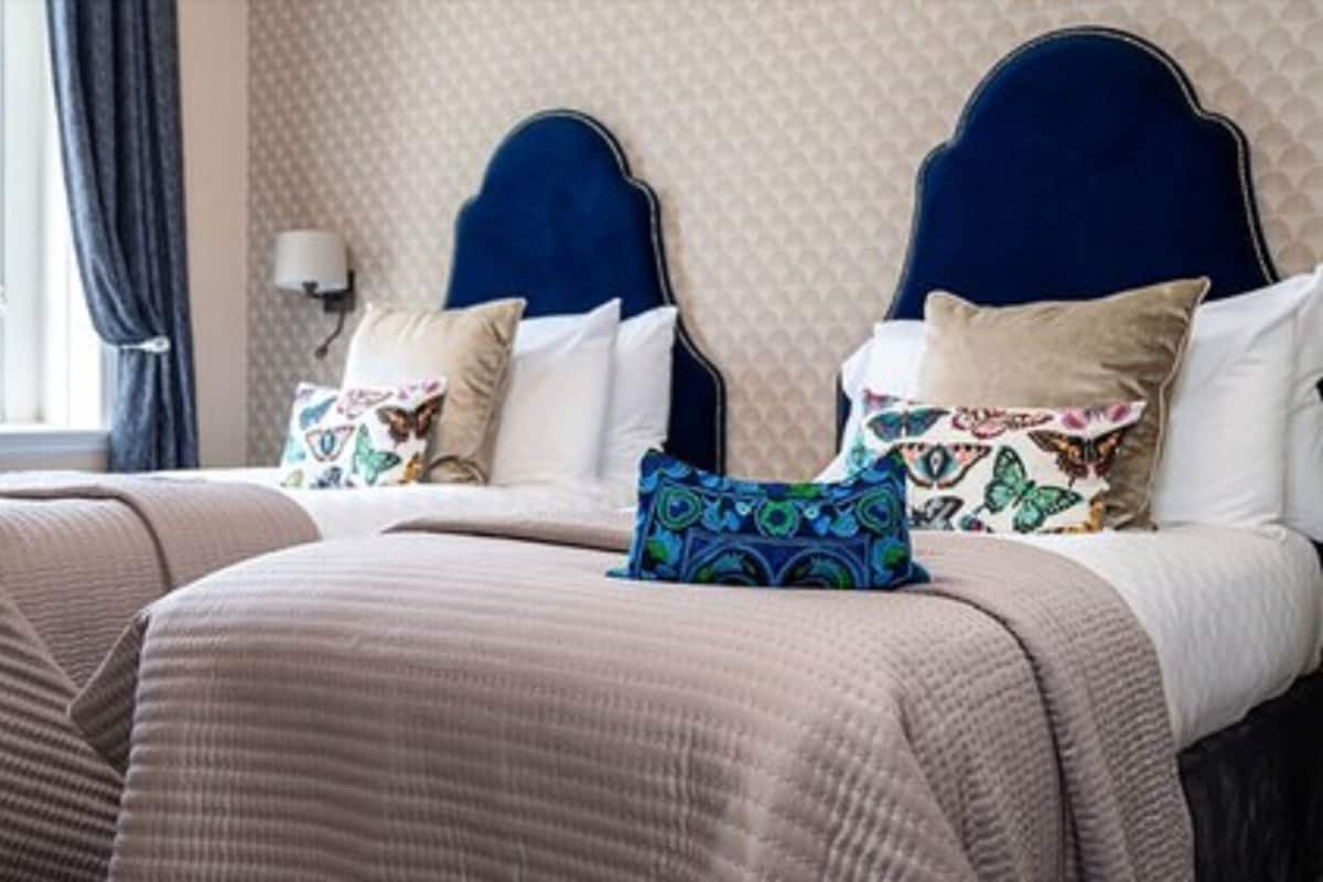 A hotel room with two beds, each with a headboard, multiple pillows, and a window with curtains.