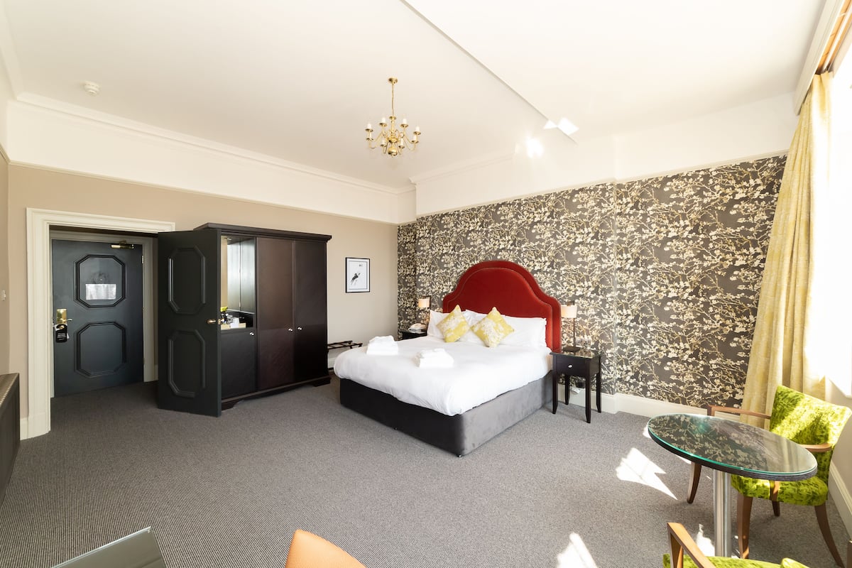 A hotel room with a large bed, a red headboard, a small table, and a chair.