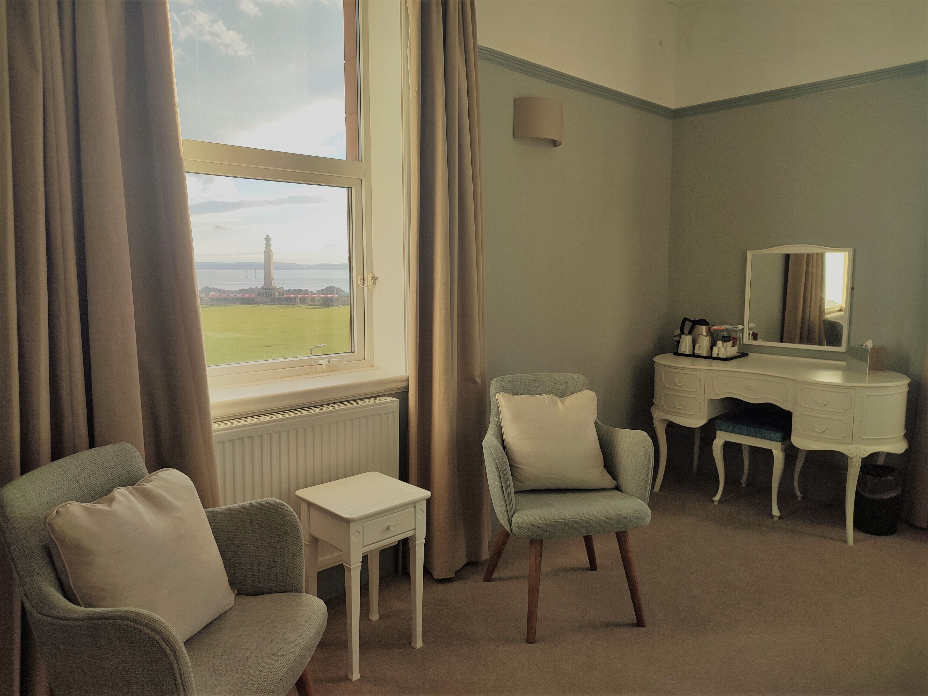 A hotel room with a view of a lighthouse, a desk with a mirror, a chair, and a small table.