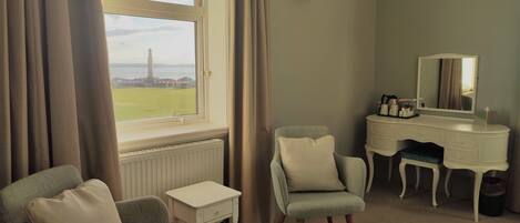 A hotel room with a view of a lighthouse, a desk with a mirror, a chair, and a small table.