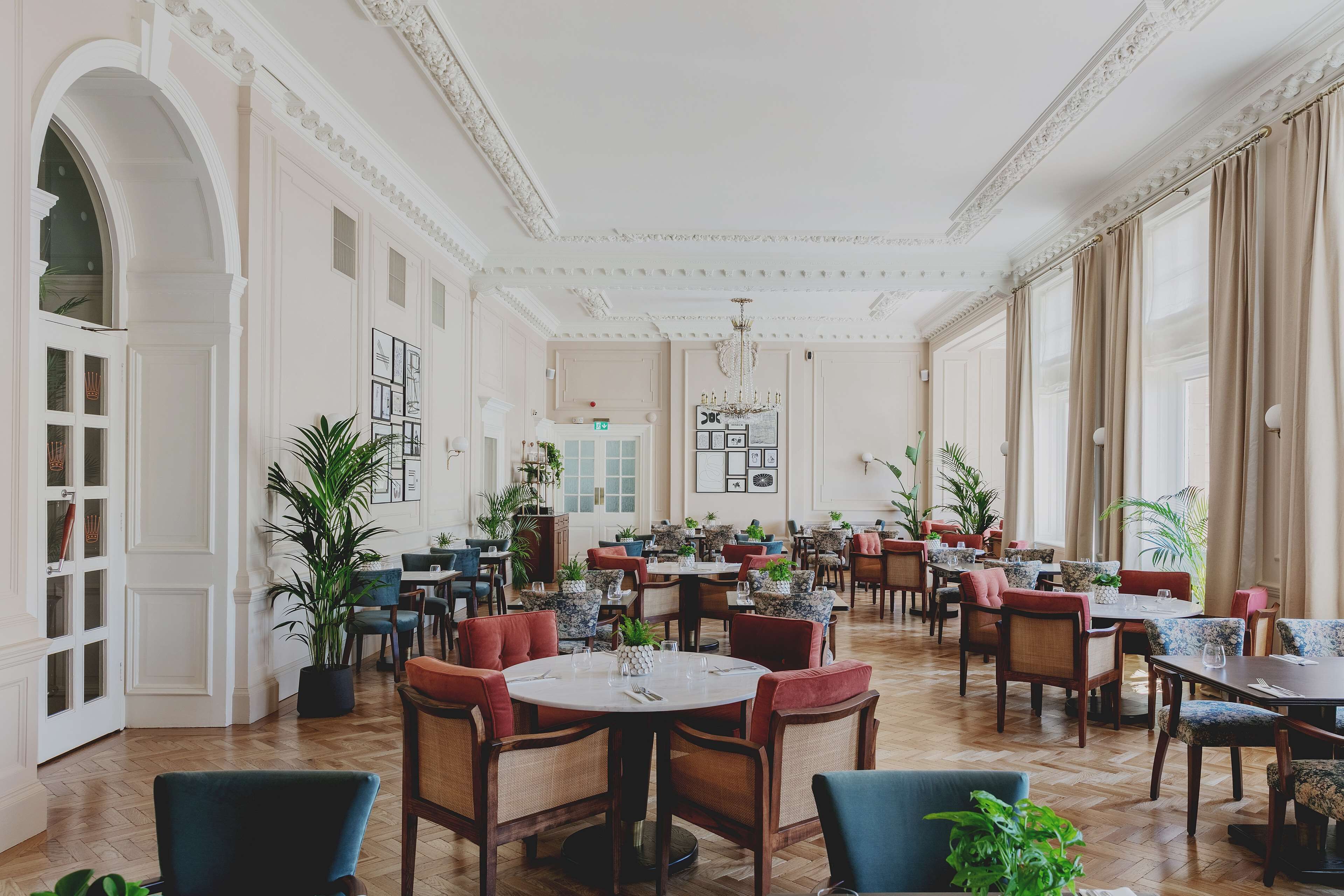 A spacious dining area with multiple tables and chairs, large windows with curtains, and potted plants.