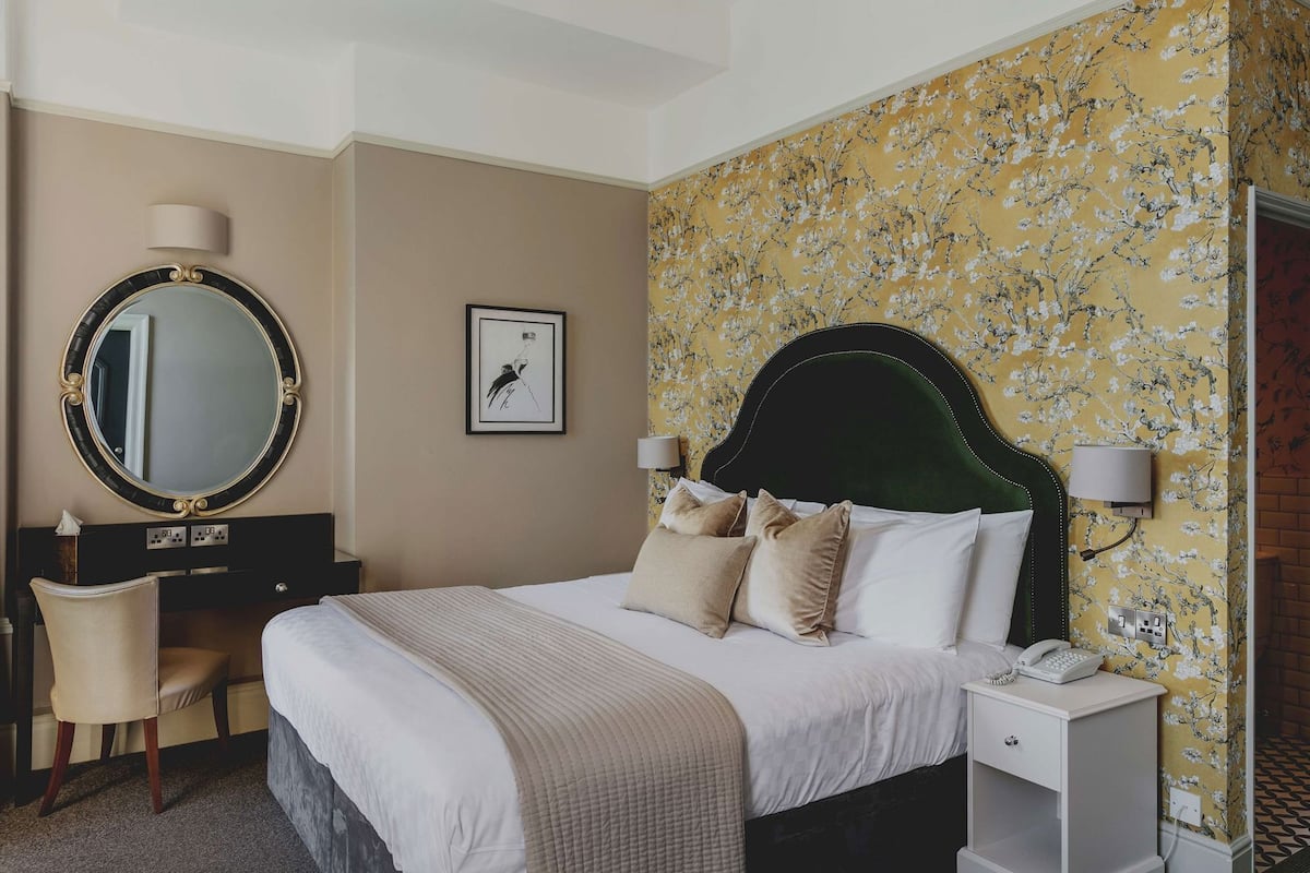 A hotel room with a large bed, a desk, a chair, a round mirror, and a framed picture on the wall.