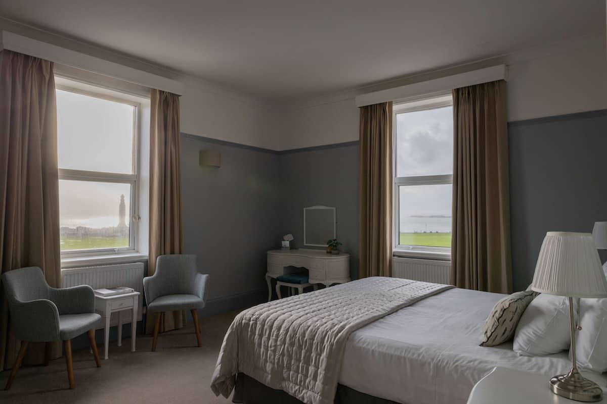 A hotel room with a bed, two chairs, a small table, a dresser, and a lamp. There are large windows with curtains, and a view of the outdoors.
