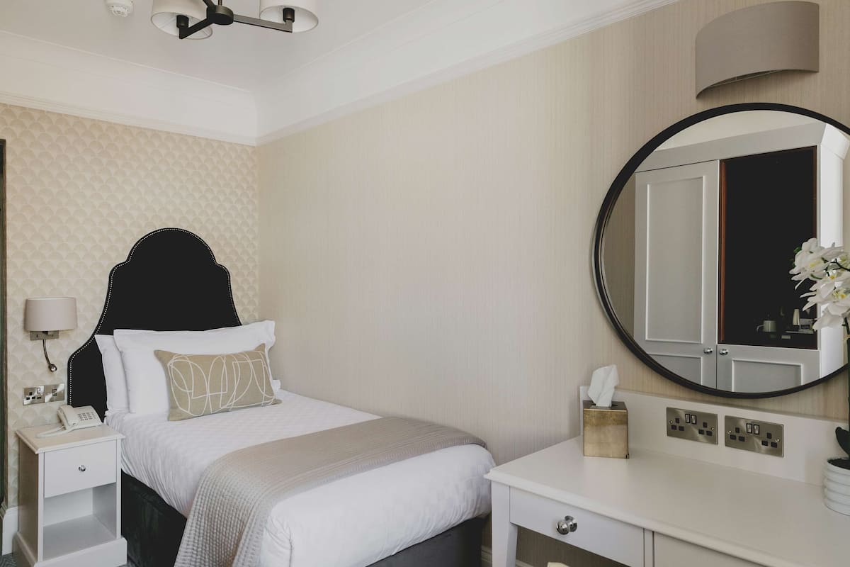 A hotel room with a bed, bedside table, lamp, mirror, and a small plant.