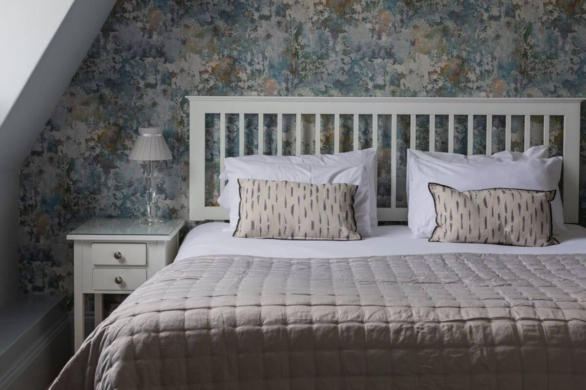 A bedroom with a sloped ceiling, a white headboard, a bed with a quilted bedspread, two pillows, a bedside table with a lamp, and a wall with a distressed floral pattern.