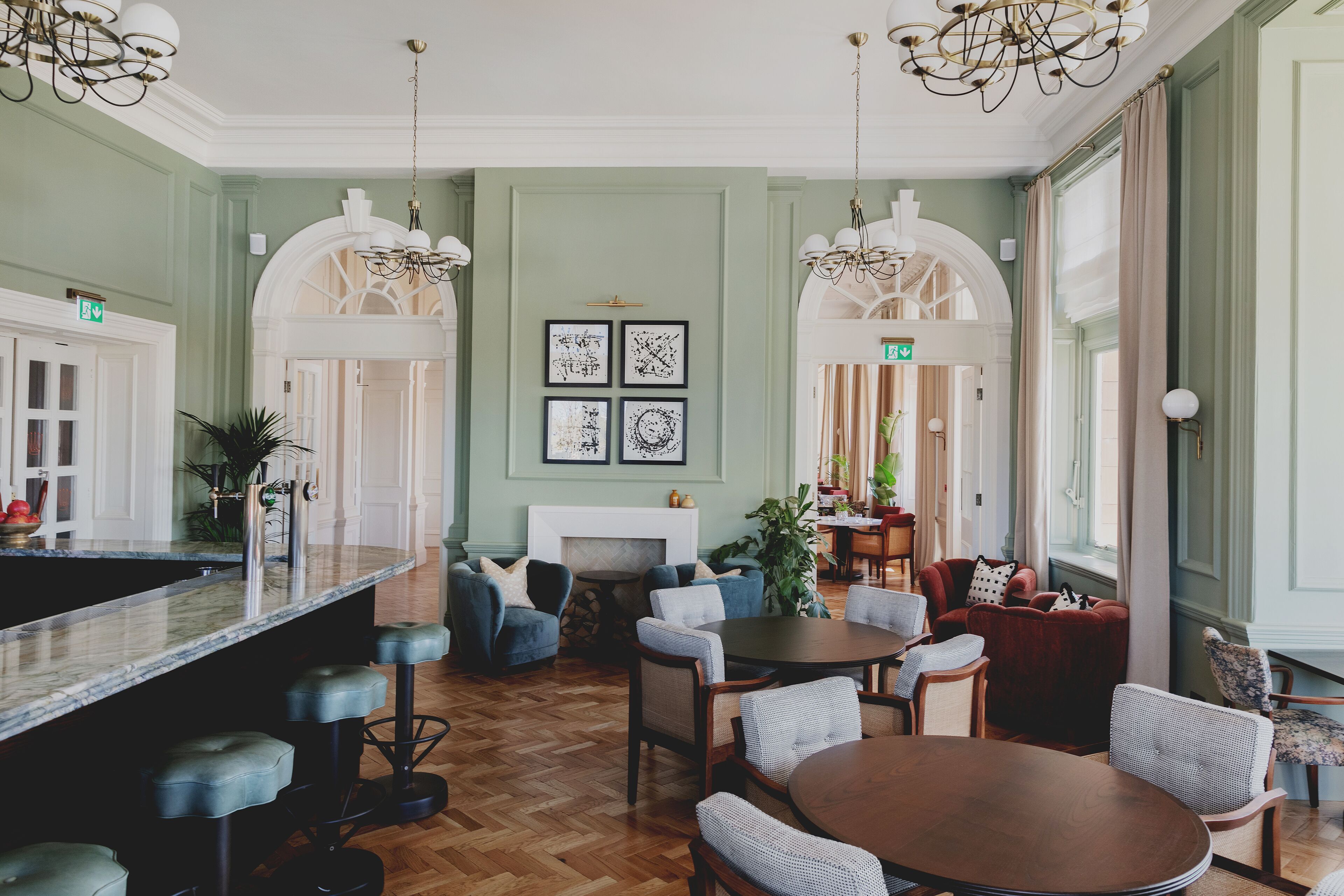 Un salon spacieux doté d’un parquet en bois à motifs chevrons, d’une cheminée centrale et d’un coin bar avec des tabourets.