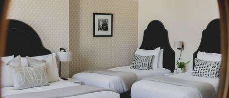 A hotel room with three beds, each with a black headboard and white bedding. There is a framed picture on the wall, a bedside lamp, and a small potted plant on one of the beds.