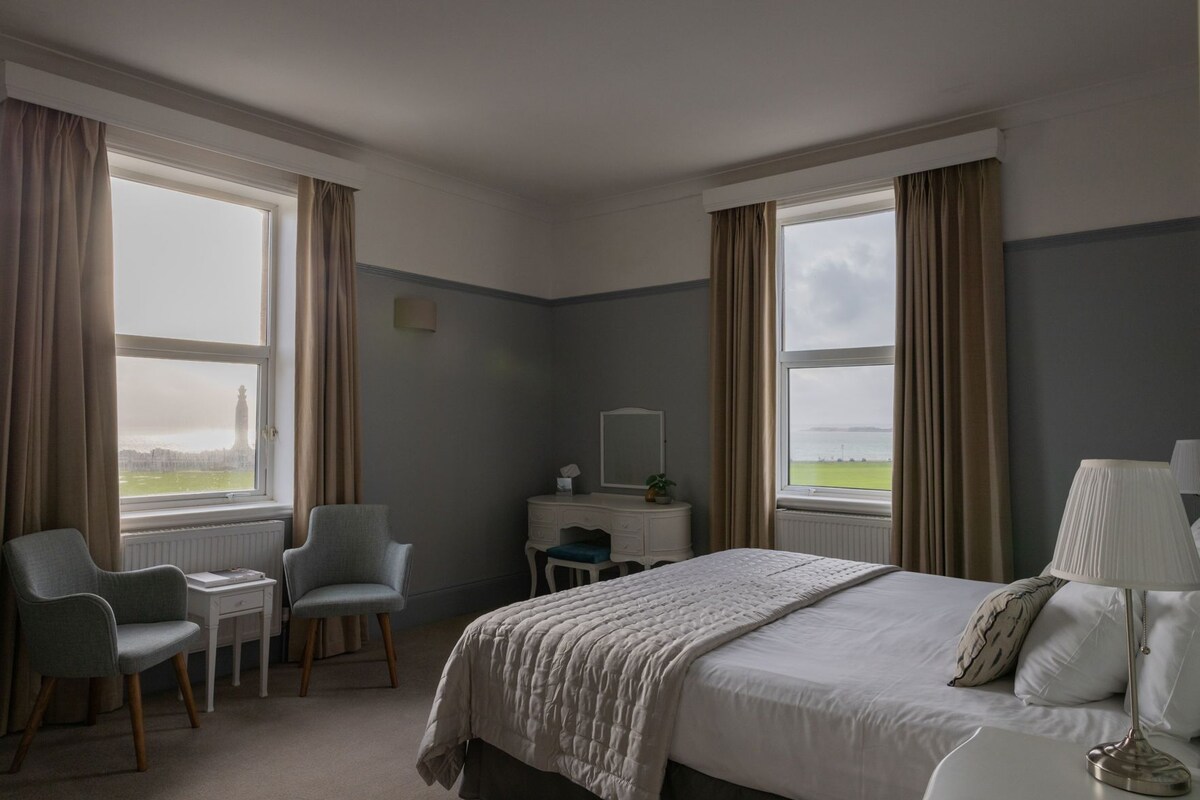 A hotel room with a bed, two chairs, a desk, a lamp, and large windows with curtains.